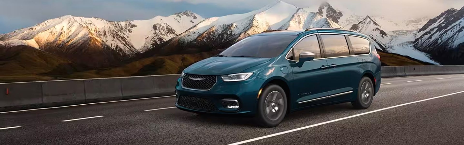 Teal minivan driving on a road alongside snow-capped mountains on a clear day.