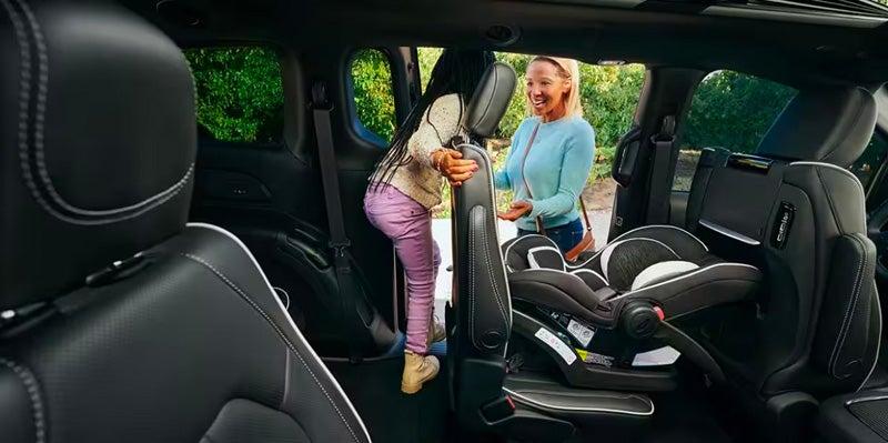 A child in pink pants and a woman smiling while leaning into a minivan or SUV with a car seat installed.