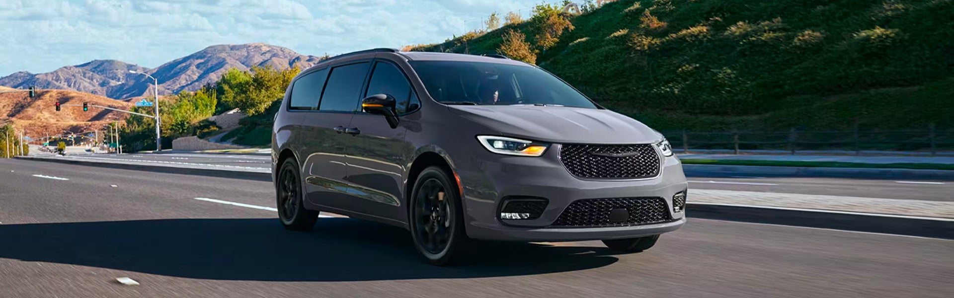 A dark gray Chrysler Pacifica Hybrid minivan drives on a multi-lane highway with mountains in the background.