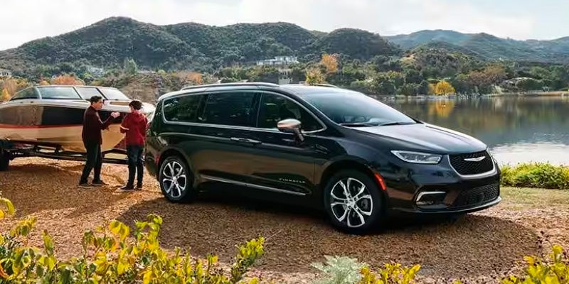 Dark minivan parked near a lake with small mountain background as two people stand by a boat on a trailer.