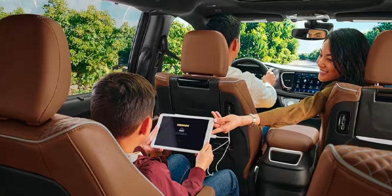 Smiling woman in passenger seat reaches back to adjust a tablet for a child in the car's second row.