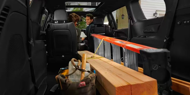Car interior from inside the trunk, showing a ladder, lumber, and tools, with a man smiling from a front seat.