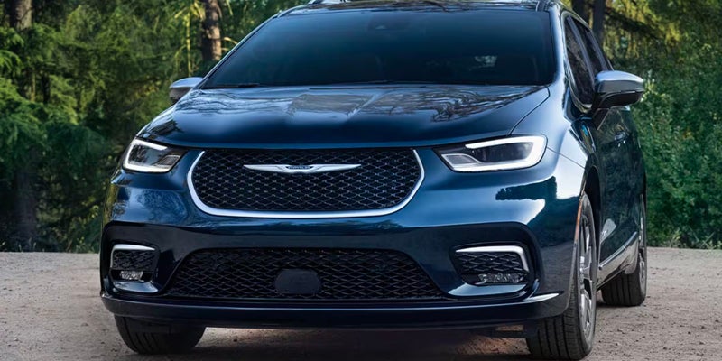 Front view of a dark blue Chrysler Pacifica minivan with bright LED headlights and a mesh grille in a natural setting.