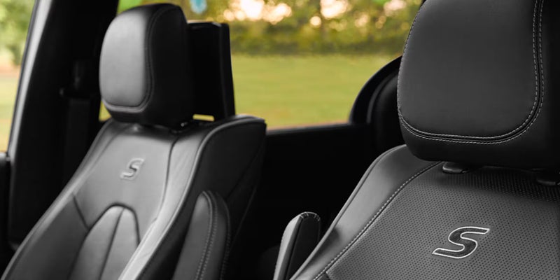 Two black leather car seats with "S" embroidered on the headrests, soft blurred green and tan background.