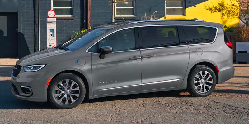 A grey Chrysler Pacifica minivan, "Pinnacle" trim, parked on a cracked asphalt street by a dark building.