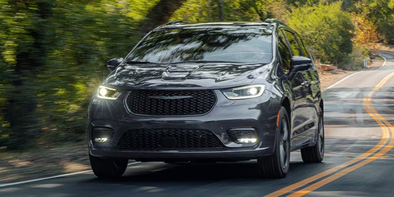 Front view of a dark gray Chrysler Pacifica minivan with bright headlights driving on a winding paved road.