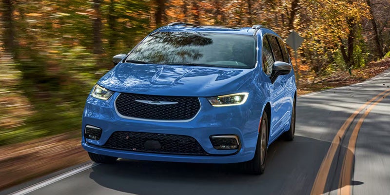 A blue minivan drives towards the viewer on a winding road bordered by autumn trees blurred by motion.