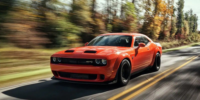 An orange Dodge Challenger Hellcat SRT drives quickly down a road lined with blurred autumn trees.
