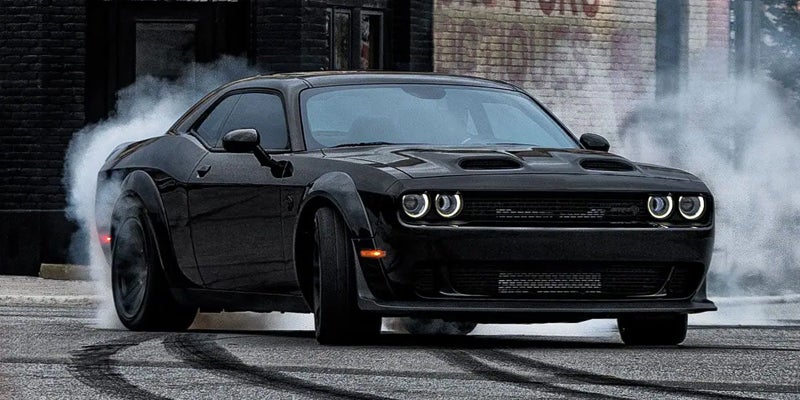 Black Dodge Challenger with smoking tires spinning on asphalt, leaving skid marks, against a brick wall.