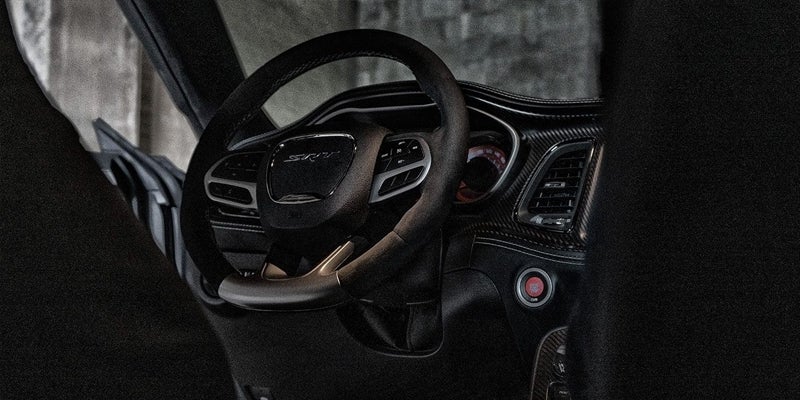 Interior view of a modern car, showing a dark steering wheel with an 'SRT' logo, dashboard, and red start button.