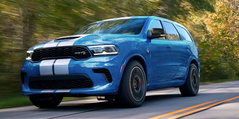 Front 3/4 view of a blue Dodge Durango SUV with white racing stripes driving on a road, trees blurring in the background.