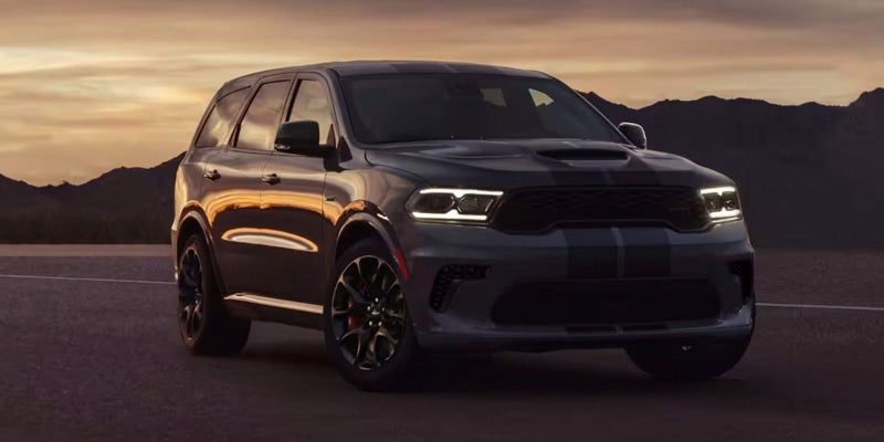 Dark gray Dodge Durango SRT Hellcat SUV with white headlights, parallel stripes, on a road with mountains at dusk.