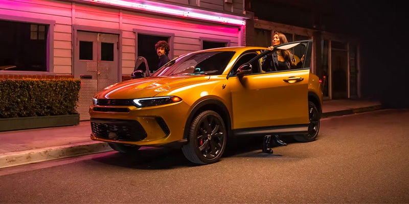 A vibrant gold Dodge Hornet SUV parked at night with a woman stepping out, and a man looking out from inside.
