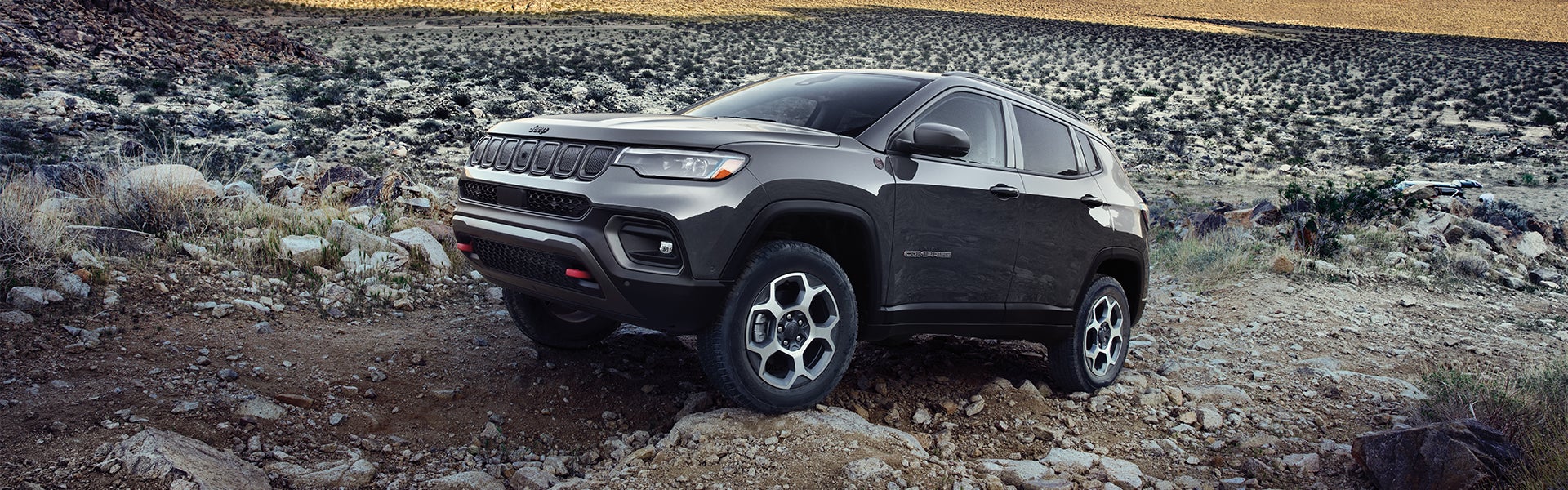 A gray Jeep Compass SUV with black trim parked on a rocky desert trail with dry brush and hills.
