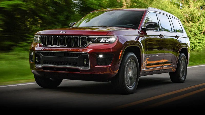 A shiny burgundy Jeep Grand Cherokee drives toward the viewer on a dark, tree-lined road.