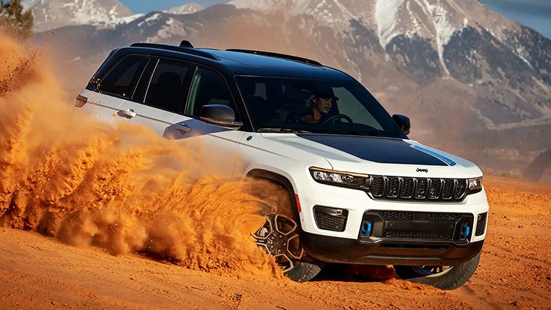 White and black Jeep Grand Cherokee kicking up red sand while driving off-road with mountains in the background.