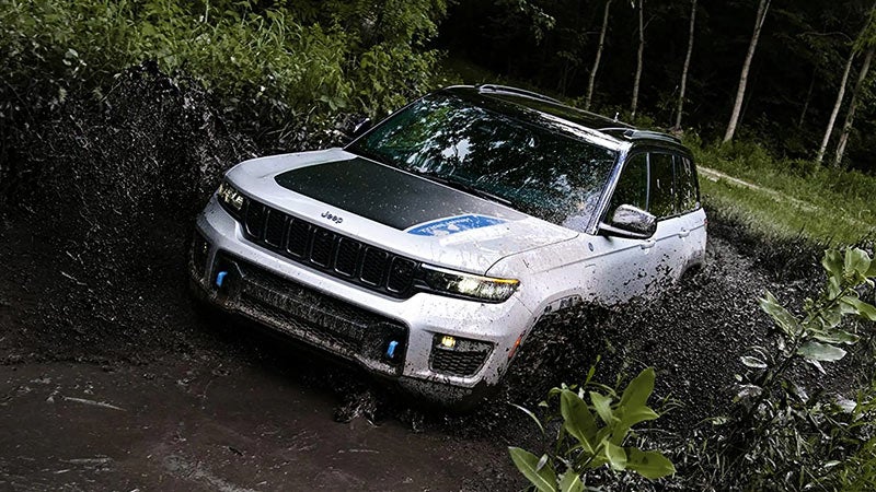 White Jeep Grand Cherokee 4xe with a black hood and blue accents, partially submerged in dark mud outdoors.