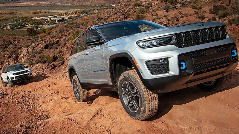 Two Jeep Grand Cherokees—silver in foreground, white in background—off-roading on a sandy, rocky path.