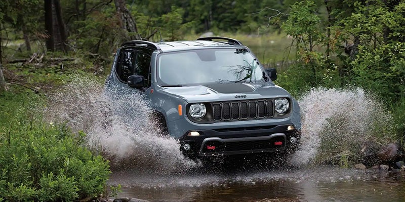 A gray Jeep Renegade splashing through a wooded stream on an overcast day.