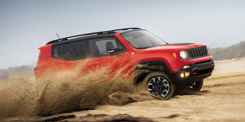 Red Jeep Renegade driving through sand, kicking up a large spray of dirt behind its wheels.