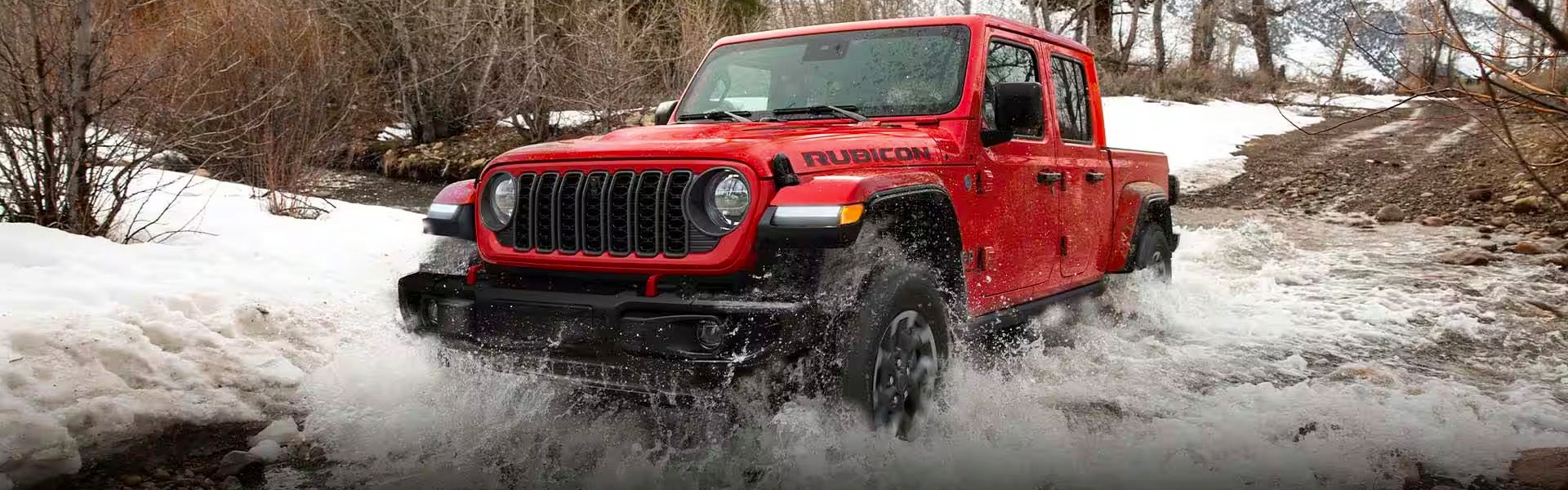 Red Jeep Rubicon off-roading through a shallow, snow-lined river, creating large splashes of water.