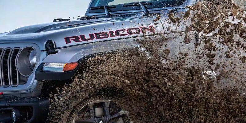 Front quarter of a gray Jeep Rubicon driving through mud, splattering it across the fender and side.