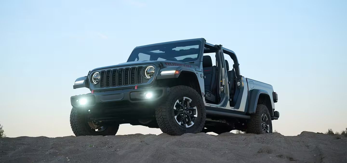 A gray Jeep Wrangler Rubicon with its doors removed, parked on a dirt hill beneath a clear sky.
