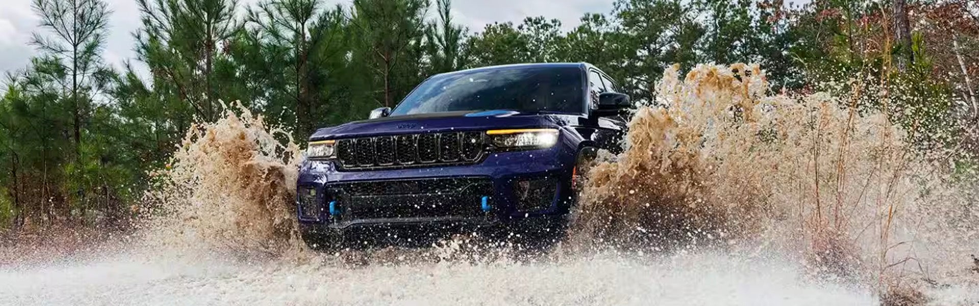 A dark blue SUV splashes through a large puddle, sending brown water high into the air with pine trees in the background.