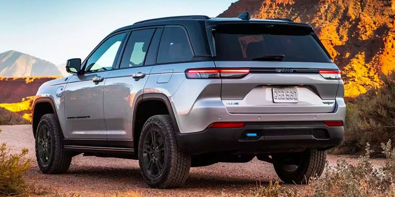 A silver Jeep Grand Cherokee 4xe with a black roof and black wheels, parked on a dirt road next to mountains.