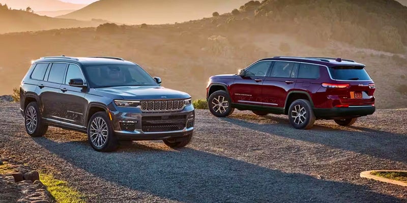 Two Jeep Grand Cherokee Ls, one dark grey and one maroon, parked on a gravel hill with mountains in the background.