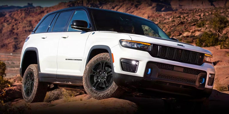 Front 3/4 view of a white Jeep Grand Cherokee off-roading on a rocky desert landscape.