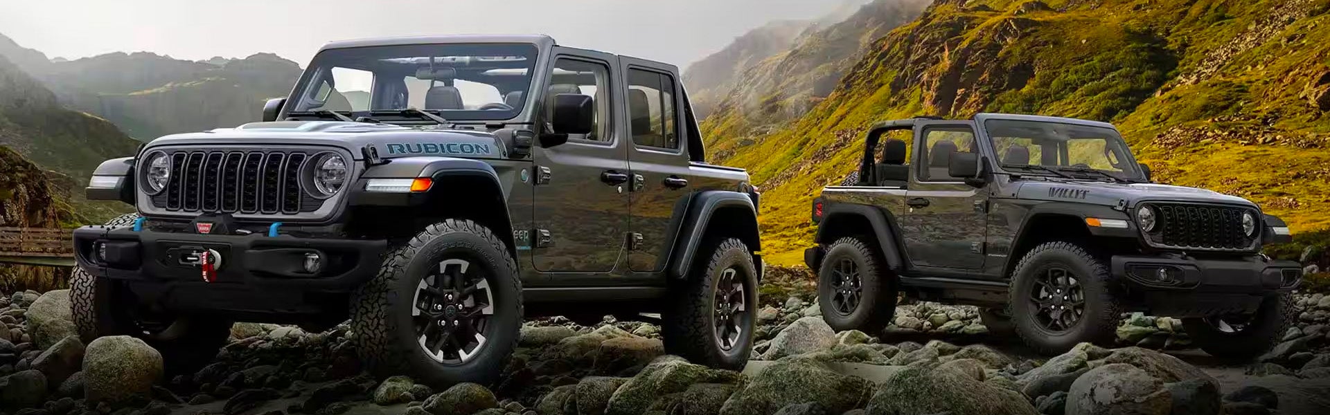 Two dark gray Jeep Wranglers, a 4-door Rubicon and a 2-door Willys, on a rocky terrain with mountains.