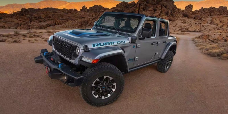 Gray Jeep Wrangler Rubicon 4xe with blue accents on a dirt road in a rocky, mountainous desert landscape at dusk.