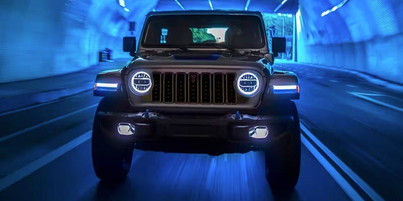 Front-facing view of a dark Jeep Wrangler with glowing headlights driving in a blue illuminated tunnel.