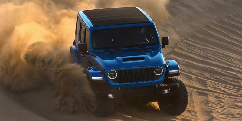 Blue Jeep Wrangler kicking up a large cloud of sand as it accelerates up a dune.