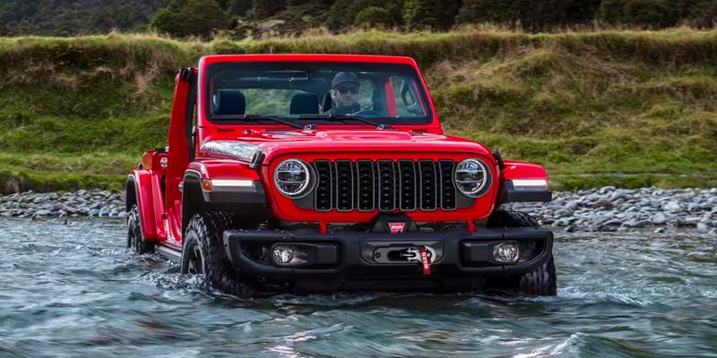 A red Jeep Wrangler Rubicon fording through a shallow river, with a green, hilly bank and trees in the background.