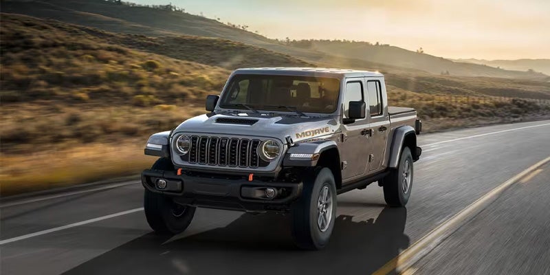Silver Jeep Gladiator Mojave pickup truck driving on an asphalt road through a desert landscape at sunset.
