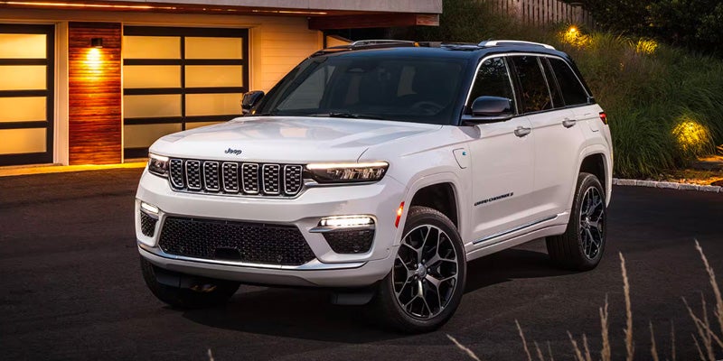 A white 2024 Jeep Grand Cherokee Summit 4xe parked on asphalt in front of a modern garage at twilight.