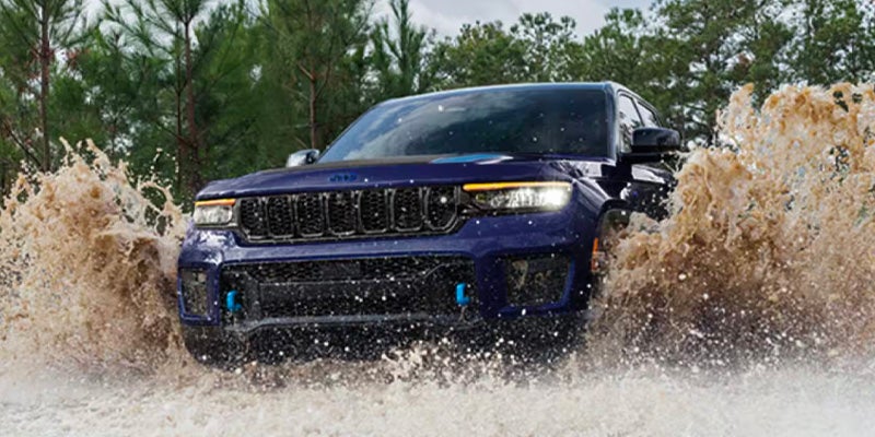 A dark blue Jeep Grand Cherokee 4xe splashes through muddy water, with trees and cloudy sky in the background.