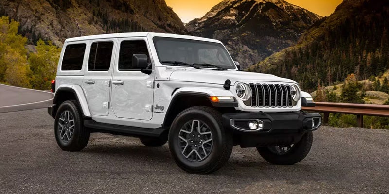 White Jeep Wrangler parked on an asphalt road with mountains and forest in the background at sunset.