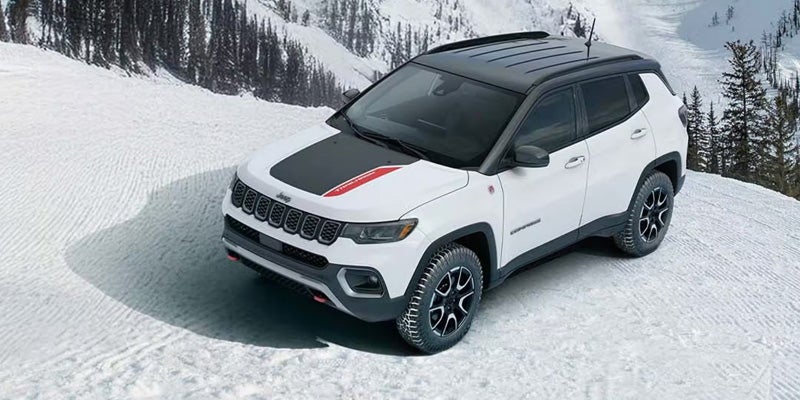 White Jeep Compass with a black roof and hood stripe, parked diagonally on a snowy ski slope.