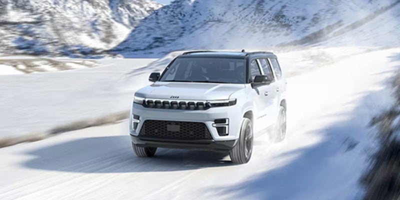 A white Jeep Wagoneer SUV driving on a snowy mountain road, with snow-covered slopes in the background.