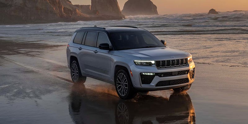 A silver 2021 Jeep Grand Cherokee L driving on a sandy beach next to the ocean at sunset.