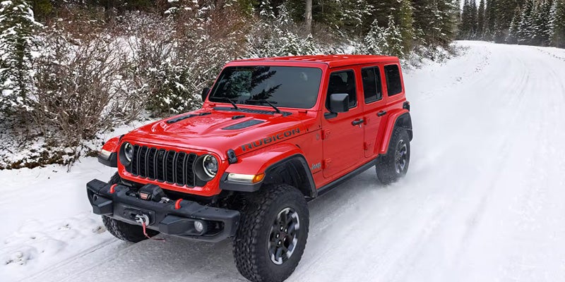 Red Jeep Rubicon 4x4 driving along a snowy road with pine trees in the background.