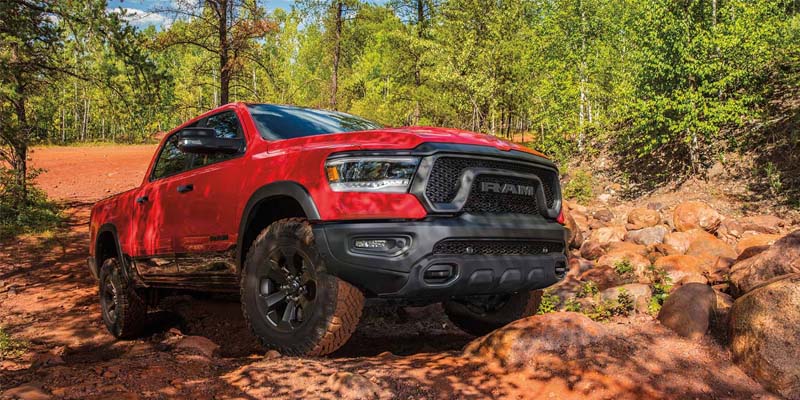 Red Ram 1500 truck with black wheels and aggressive tires off-roading on a dirt path with large rocks.