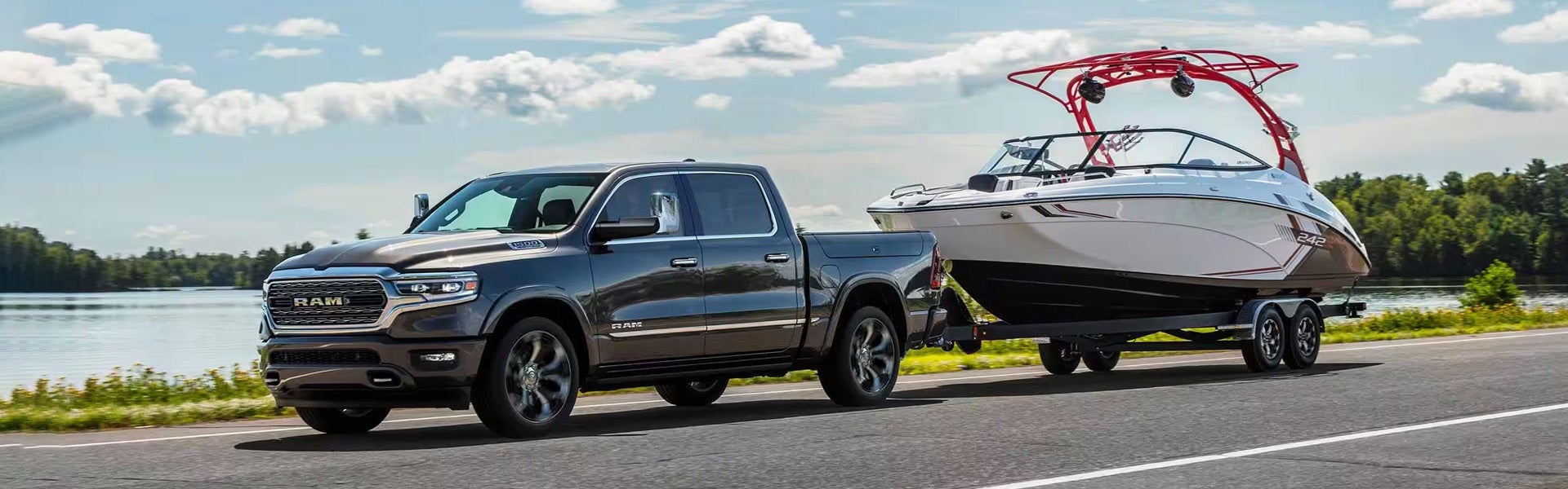 A dark grey RAM truck towing a white motorboat on a trailer, driving on an asphalt road next to a lake.