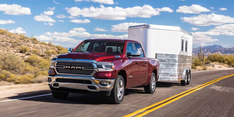 A red Ram truck towing a silver horse trailer on a desert highway with mountains and cactuses.
