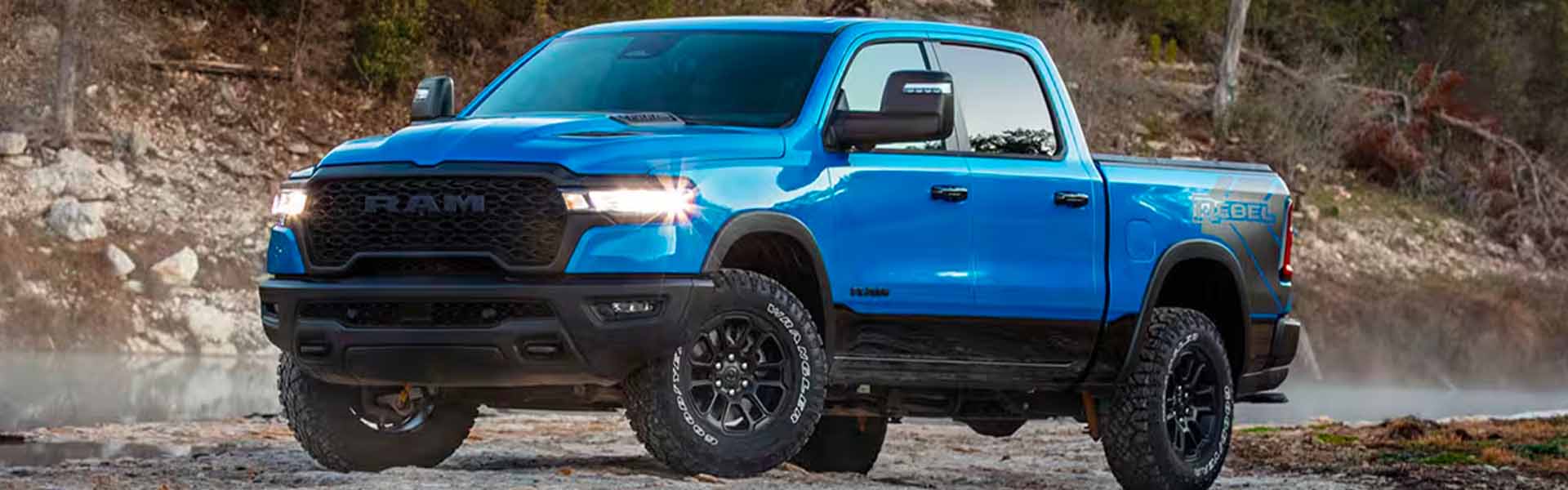 A bright blue Ram Rebel pickup truck with glowing headlights seen from a low angle, showcasing large tires on dirt and rocks with a hint of water.