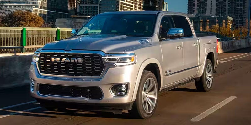 A silver 2024 Ram 1500 truck with a prominent grille drives on a road with city buildings in the background.