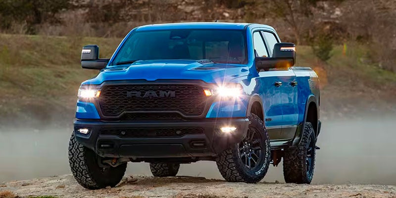 A blue Ram TRX pickup truck with its headlights on, driving on a dirt road.